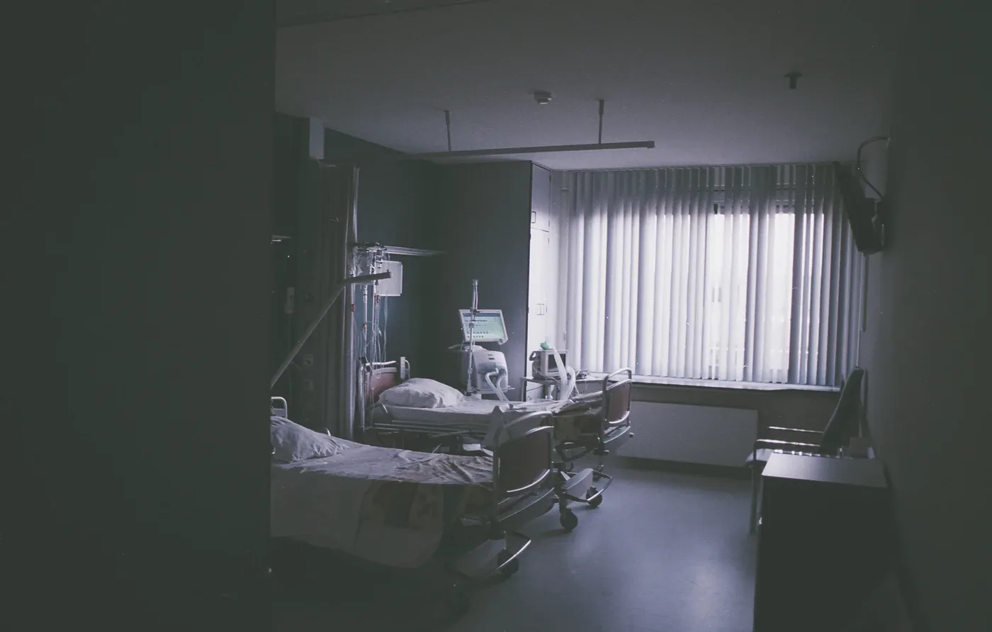An empty hospital ward with two beds, medical equipment and light coming through vertical blinds.